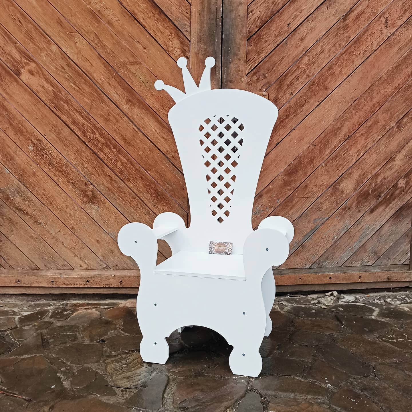 White wooden event throne chair featuring a crown top and a diamond lattice cutout on the backrest. Displayed outdoors on a stone patio against a rustic wooden door. Elegant seating prop for rustic weddings, birthdays, and garden parties.
