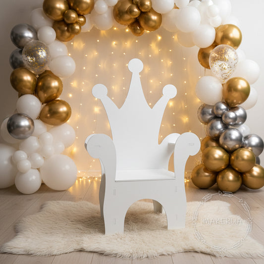 White wooden kids throne chair with a classic crown-shaped backrest featuring rounded tips. Styled indoors on a fluffy white rug in front of a white and gold balloon garland with fairy lights. Perfect royal photo prop for first birthdays and baby showers.