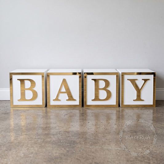 Decorative white wooden blocks with mirror gold plastic, spelling 'BABY' on a wooden floor with a white wall background.